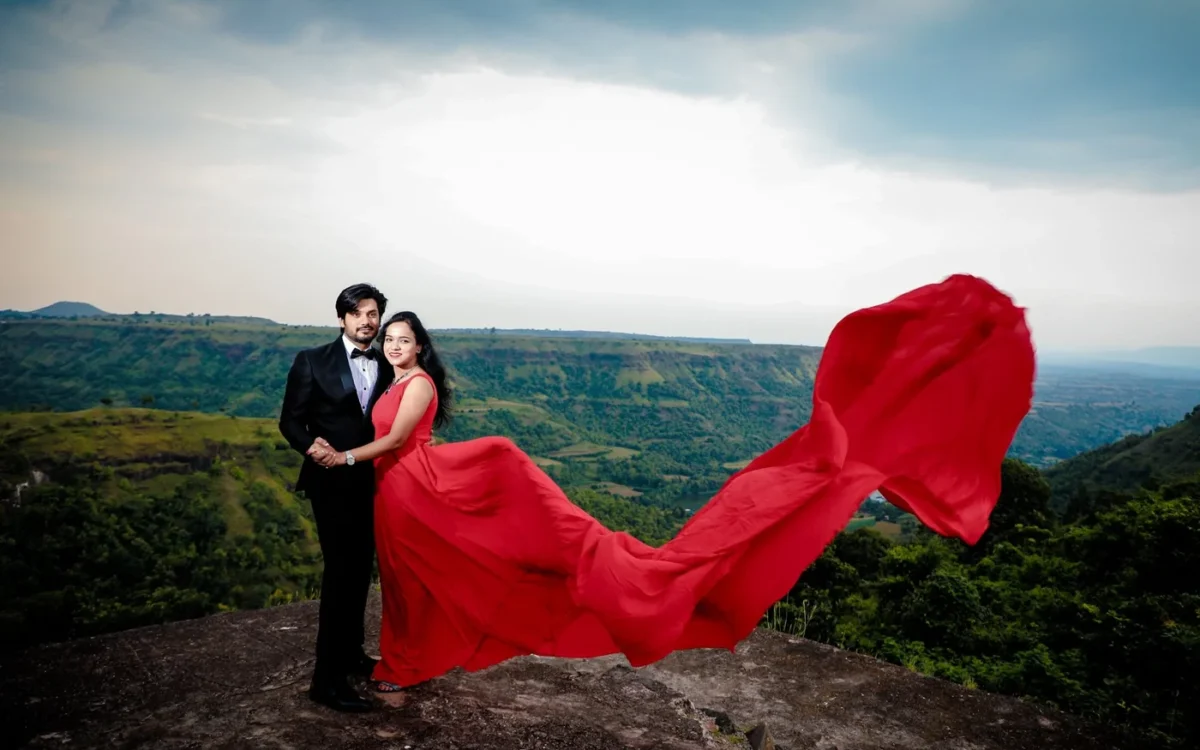 Couple in a red flowing gown and black tuxedo standing on a rocky hilltop viewpoint above green valleys, pre-wedding shoot near Indore