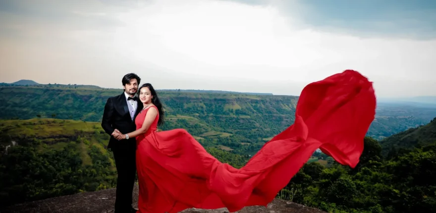 Couple in a red flowing gown and black tuxedo standing on a rocky hilltop viewpoint above green valleys, pre-wedding shoot near Indore