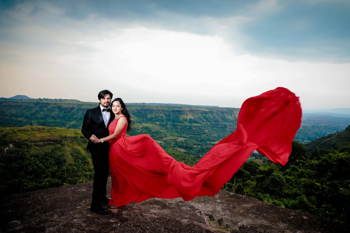 Couple in a red flowing gown and black tuxedo standing on a rocky hilltop viewpoint above green valleys, pre-wedding shoot near Indore