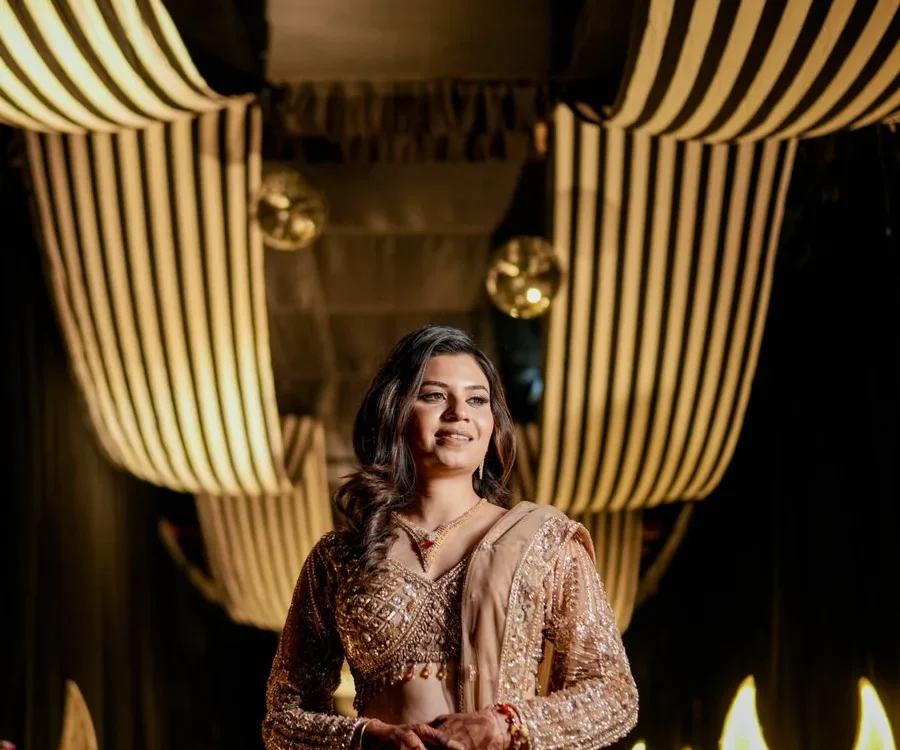 Bride in an embroidered gold lehenga posing for bridal portraits under a draped ceiling with warm lights