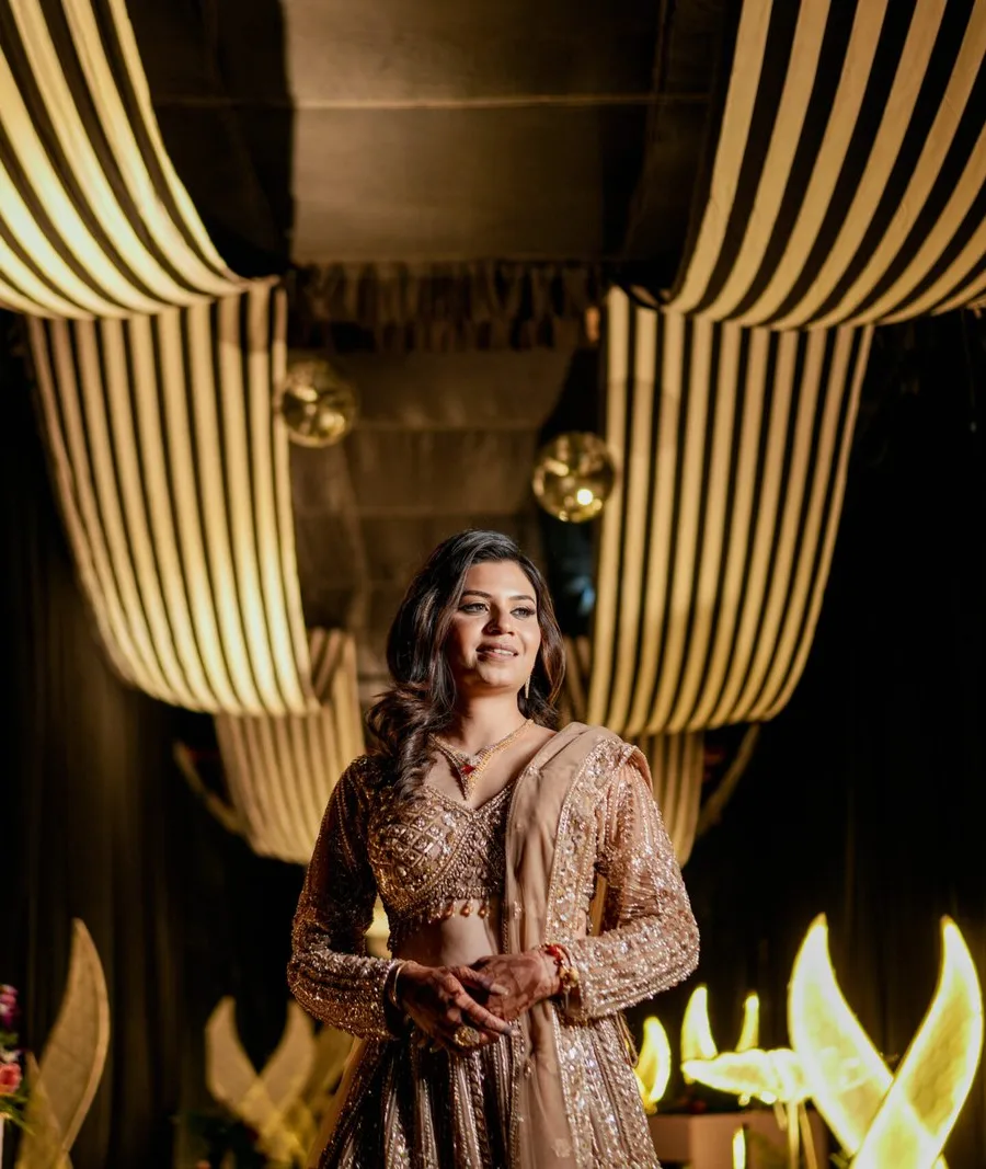 Bride in an embroidered gold lehenga posing for bridal portraits under a draped ceiling with warm lights