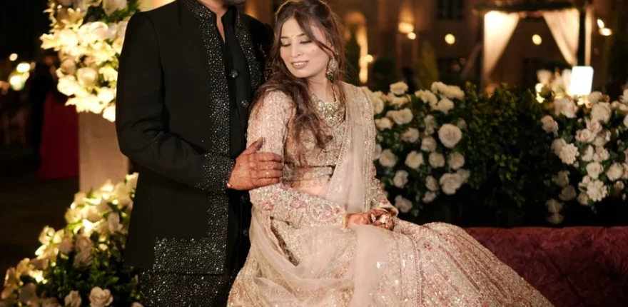 Bride in an embellished ivory lehenga seated beside the groom in a black sequinned sherwani under floral chandeliers at an elegant wedding reception
