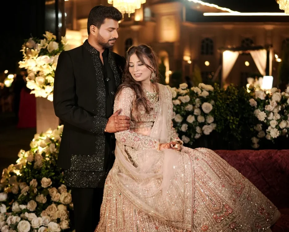 Bride in an embellished ivory lehenga seated beside the groom in a black sequinned sherwani under floral chandeliers at an elegant wedding reception