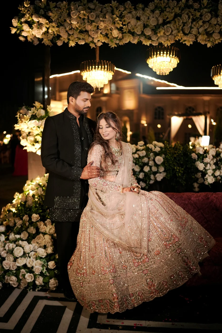 Bride in an embellished ivory lehenga seated beside the groom in a black sequinned sherwani under floral chandeliers at an elegant wedding reception