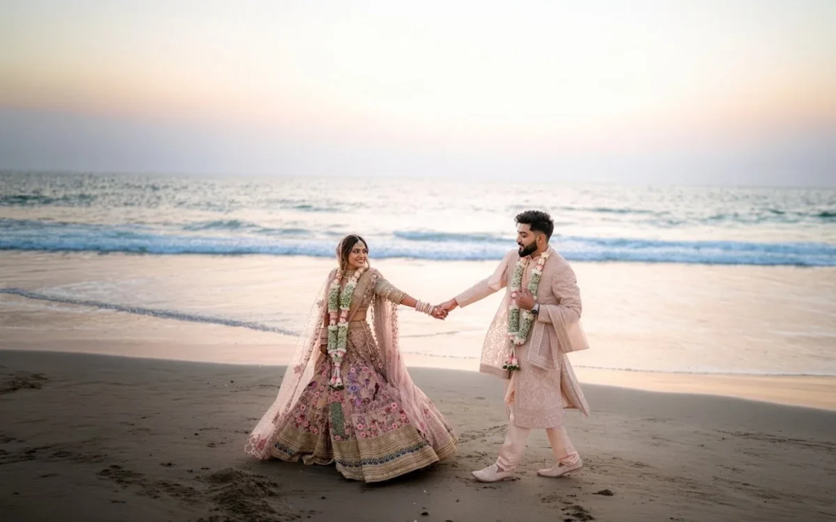 Bride in a pink embroidered lehenga and groom in a cream sherwani holding hands on a sandy beach at sunset