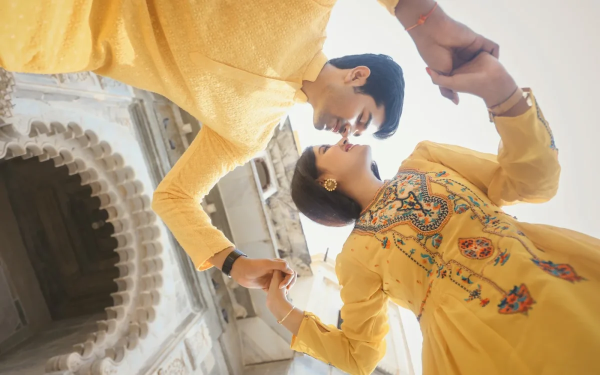 Couple in yellow outfits holding hands and leaning in close beside an arched stone wall, a haldi-style portrait in Indore
