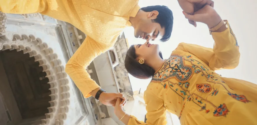 Couple in yellow outfits holding hands and leaning in close beside an arched stone wall, a haldi-style portrait in Indore