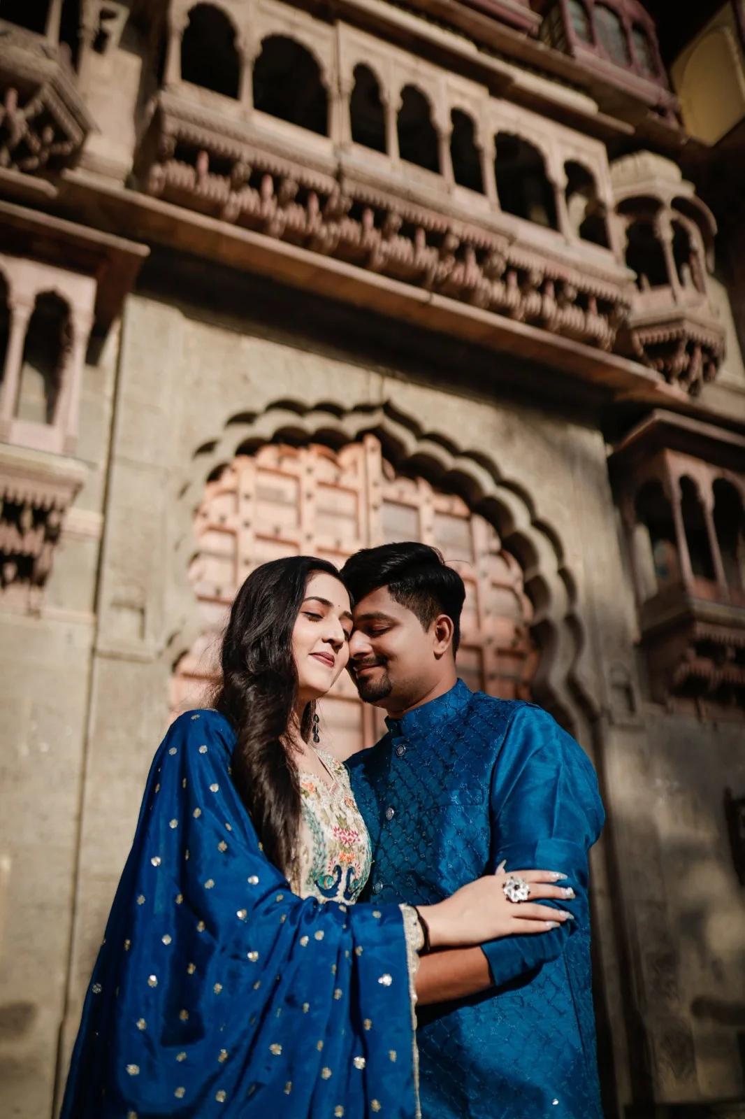 Couple in coordinated blue traditional outfits sharing a quiet moment beneath an ornate carved stone arch