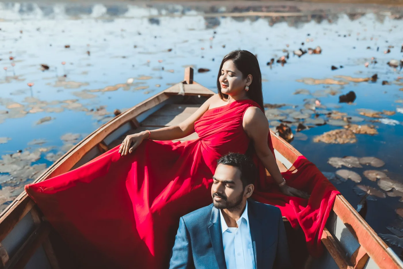 Couple in a wooden boat on a lily pond, woman in a flowing red gown and man in a navy blazer, relaxed pre-wedding shoot near Indore