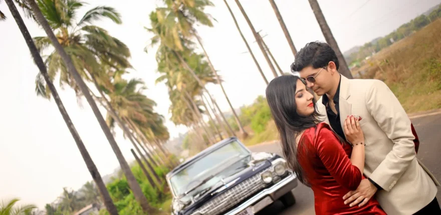 Couple in red and cream formal outfits embracing beside a classic car on a palm-lined road