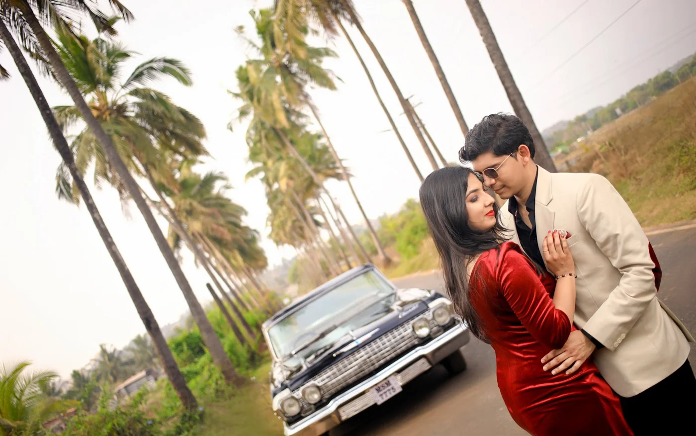 Couple in red and cream formal outfits embracing beside a classic car on a palm-lined road