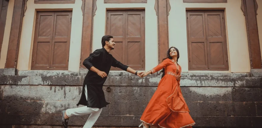 Couple running hand in hand across a heritage courtyard, woman in an orange gown and man in a black kurta, pre-wedding shoot in Indore