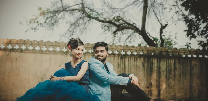 Couple sitting side by side on grass beside a stone wall, woman in a teal gown and man in a light blue blazer, pre-wedding shoot near Indore