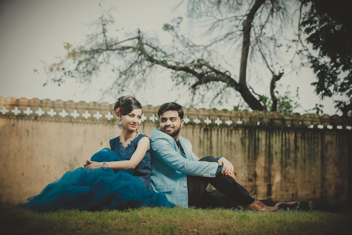 Couple sitting side by side on grass beside a stone wall, woman in a teal gown and man in a light blue blazer, pre-wedding shoot near Indore
