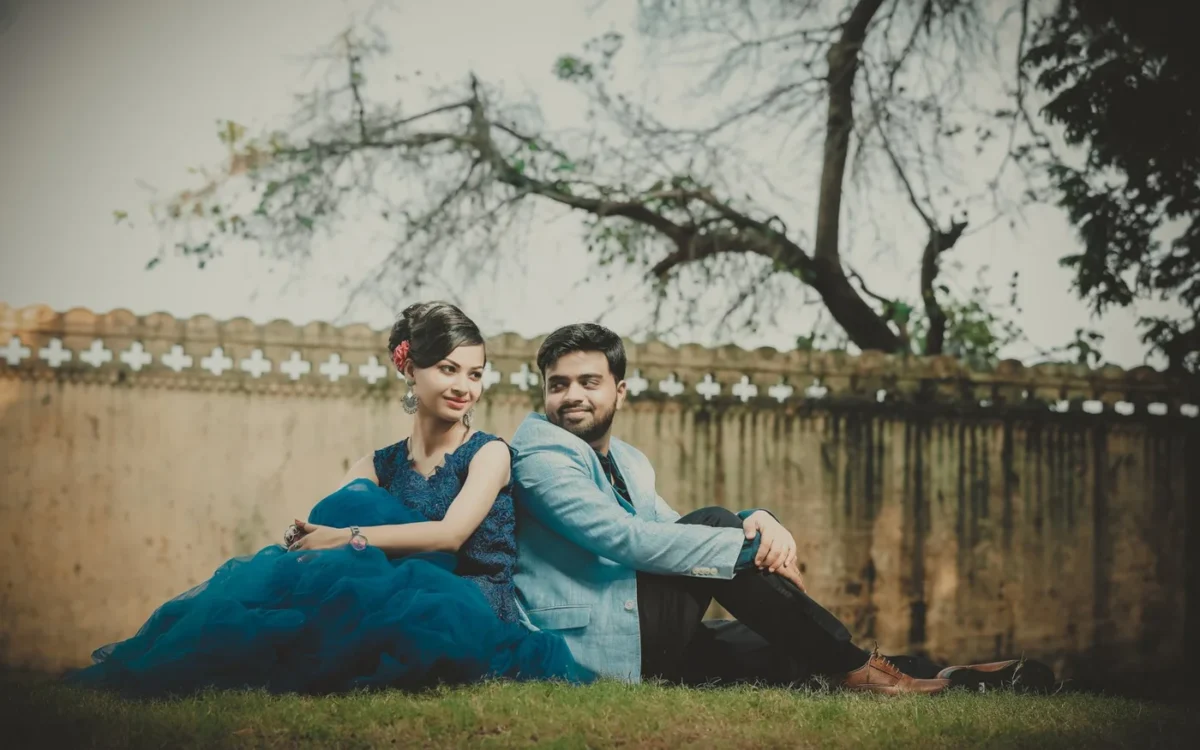 Couple in coordinated blue outfits sitting back to back on grass beside a stone wall