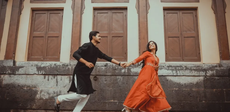 Pre-wedding shoot outfit changes - traditional couple look Couple holding hands in black and orange traditional outfits during a moving outdoor portrait