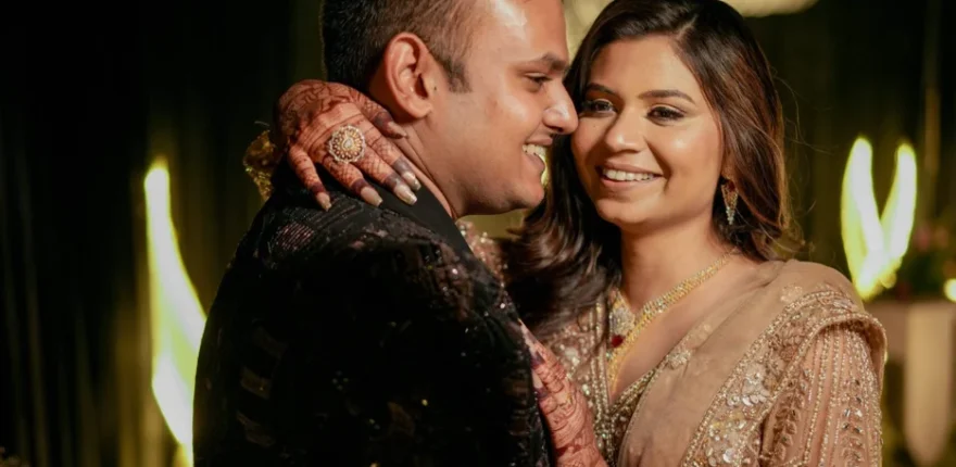 Bride in an embellished beige lehenga with mehendi on her hands embracing the groom in a black sherwani during their wedding reception, warm professional wedding photography