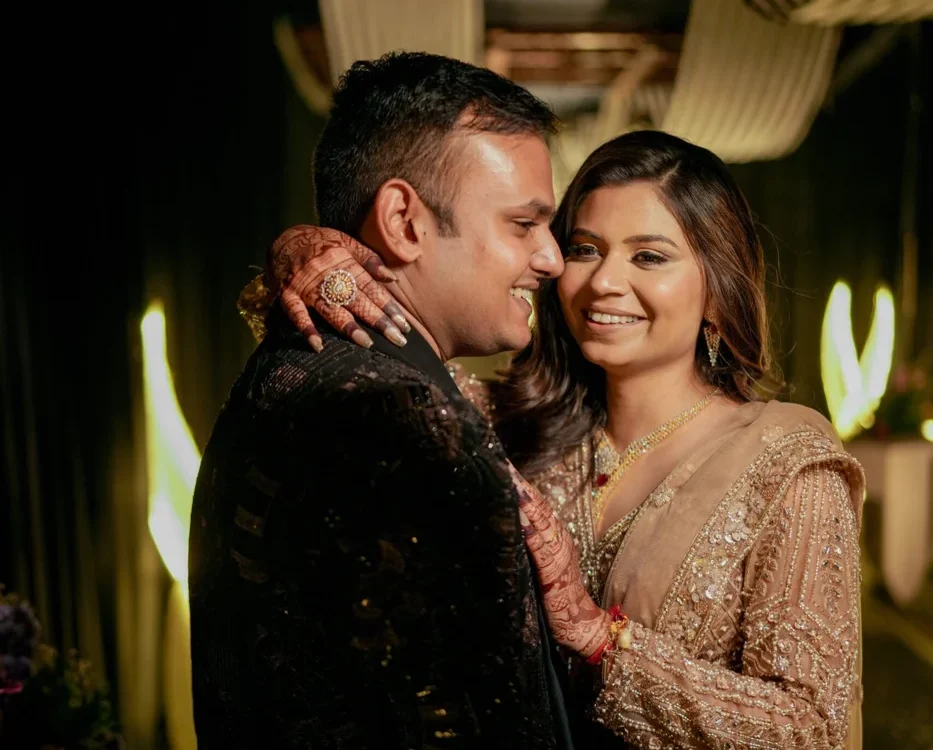 Bride in an embellished beige lehenga with mehendi on her hands embracing the groom in a black sherwani during their wedding reception, warm professional wedding photography