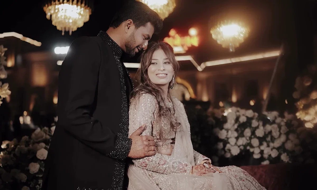 Groom in a black embellished sherwani standing beside the seated bride at their wedding reception, with chandeliers behind them