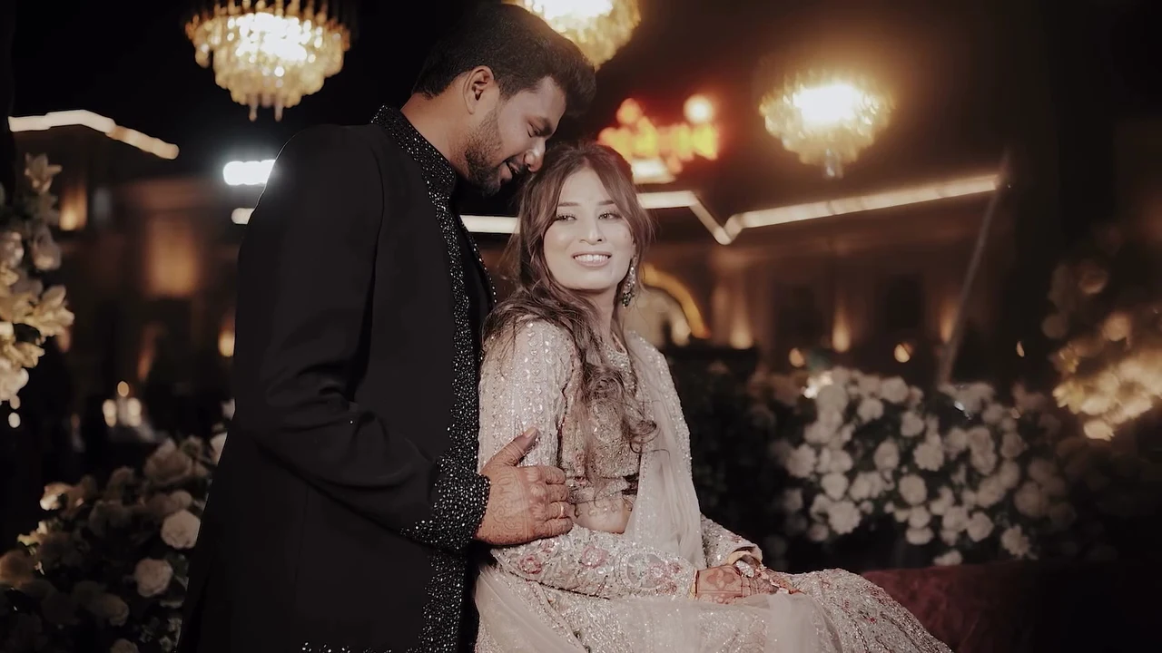 Groom in a black embellished sherwani standing beside the seated bride at their wedding reception, with chandeliers behind them