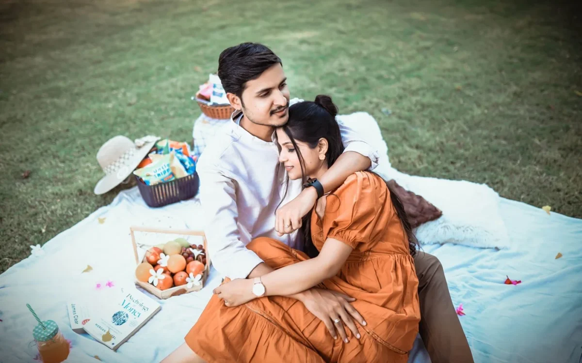 Indian couple sitting close on a picnic blanket on a lawn with fruit and a hat, a romantic pre-wedding shoot idea near Indore