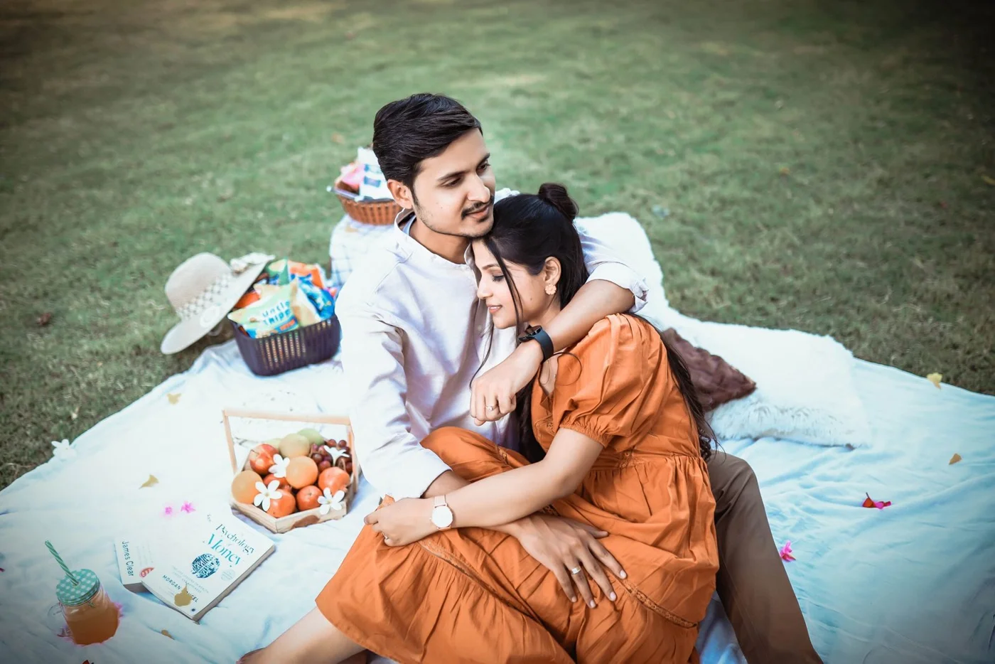 Indian couple sitting close on a picnic blanket on a lawn with fruit and a hat, a romantic pre-wedding shoot idea near Indore