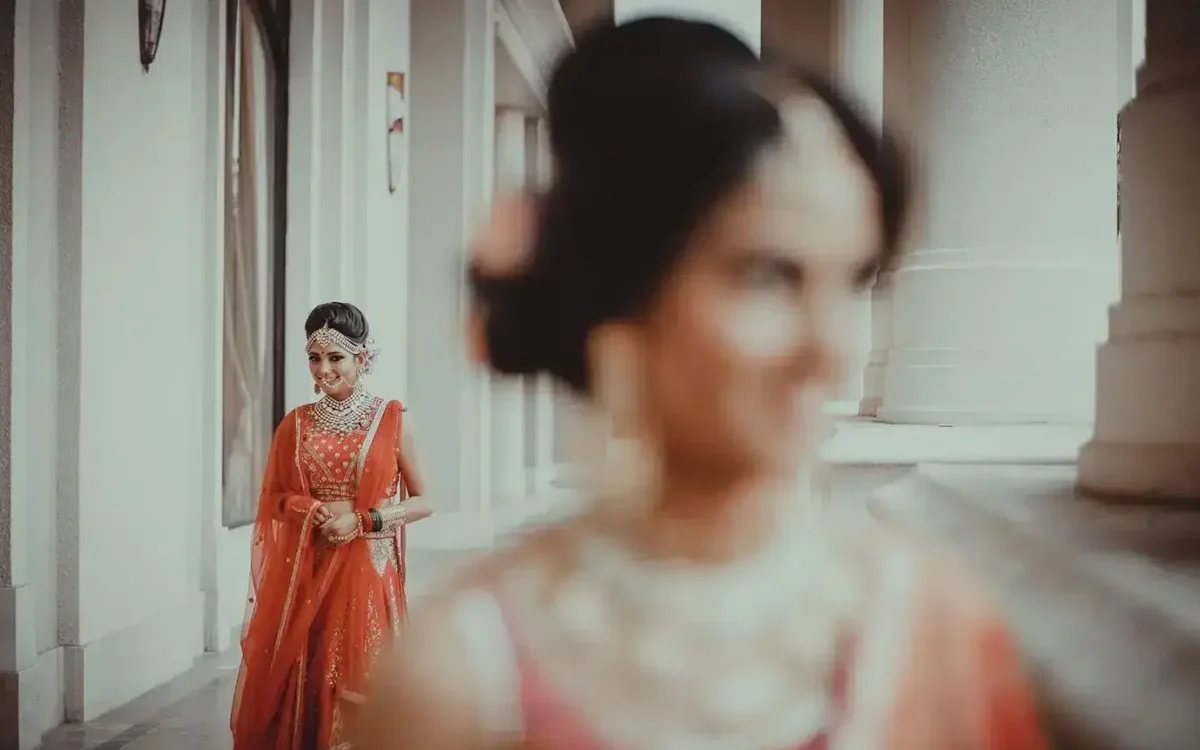 Bride in a red lehenga with kundan jewellery standing in a bright corridor, a bridesmaid softly blurred in the foreground