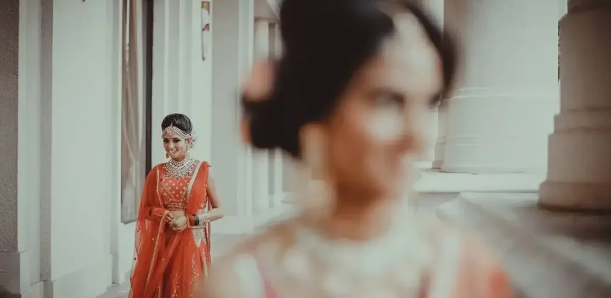 Bride in a red lehenga with kundan jewellery standing in a bright corridor, a bridesmaid softly blurred in the foreground