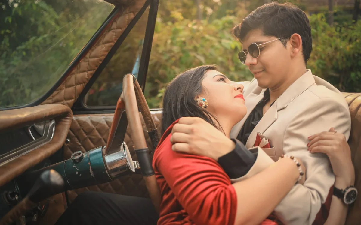 Indian couple embracing in the front seat of a vintage car, woman in red and man in a cream suit with round sunglasses, pre-wedding shoot near Indore