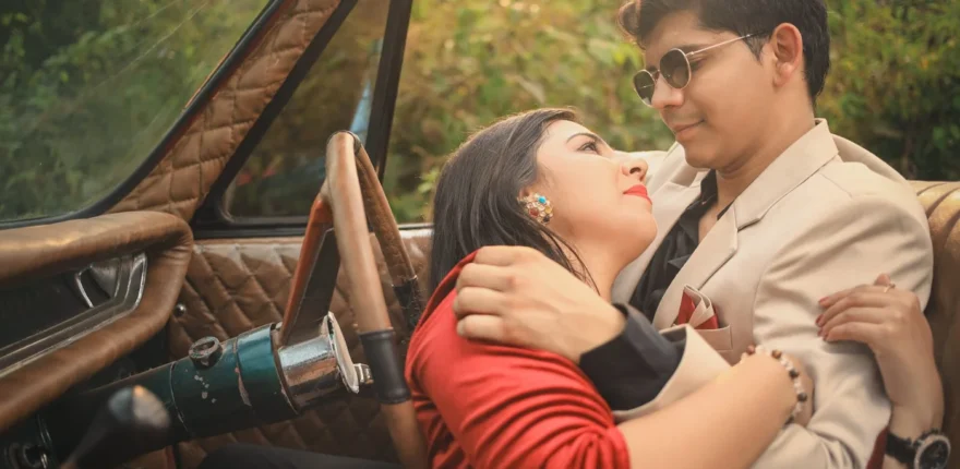 Indian couple embracing in the front seat of a vintage car, woman in red and man in a cream suit with round sunglasses, pre-wedding shoot near Indore