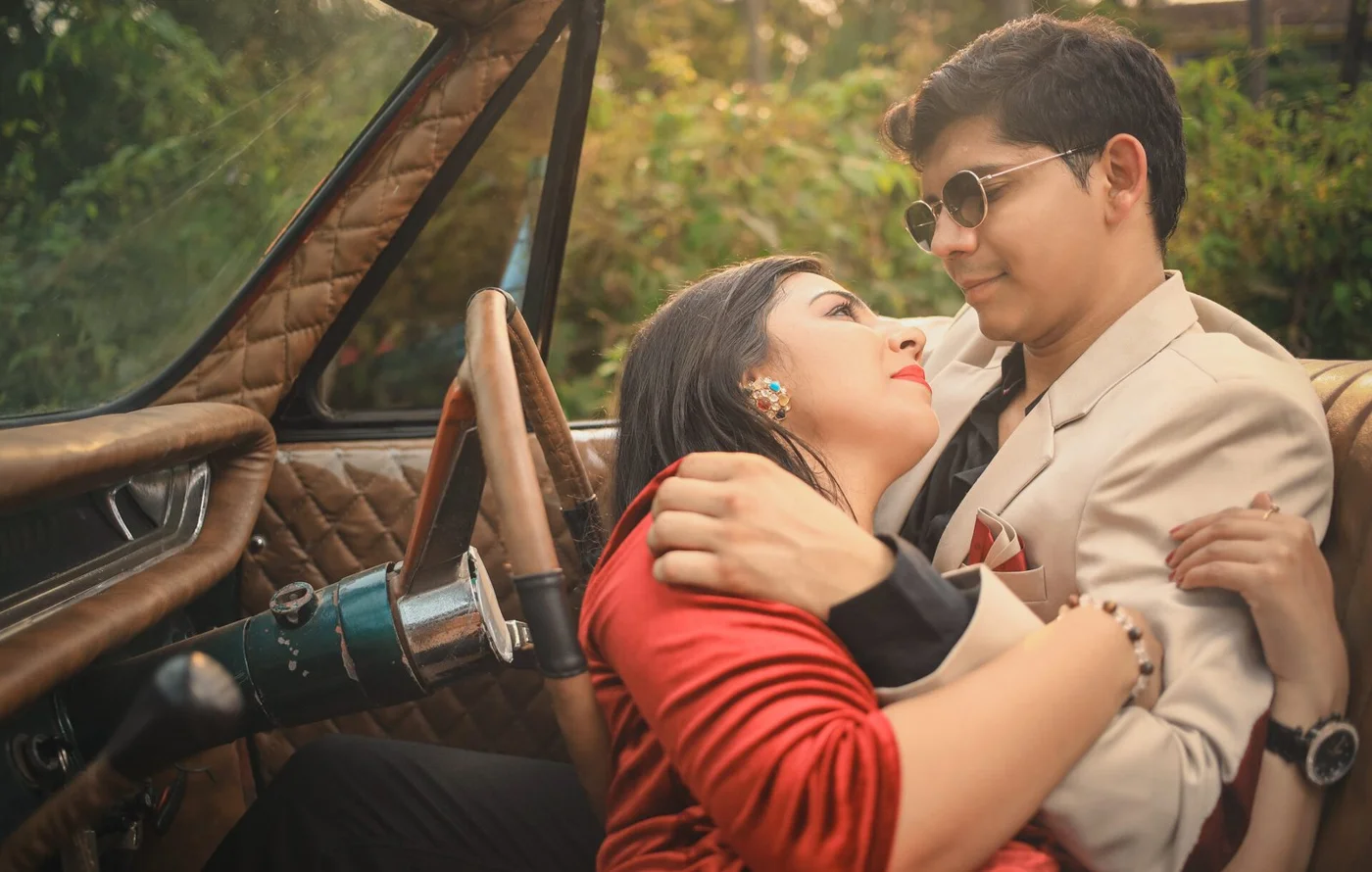 Indian couple embracing in the front seat of a vintage car, woman in red and man in a cream suit with round sunglasses, pre-wedding shoot near Indore