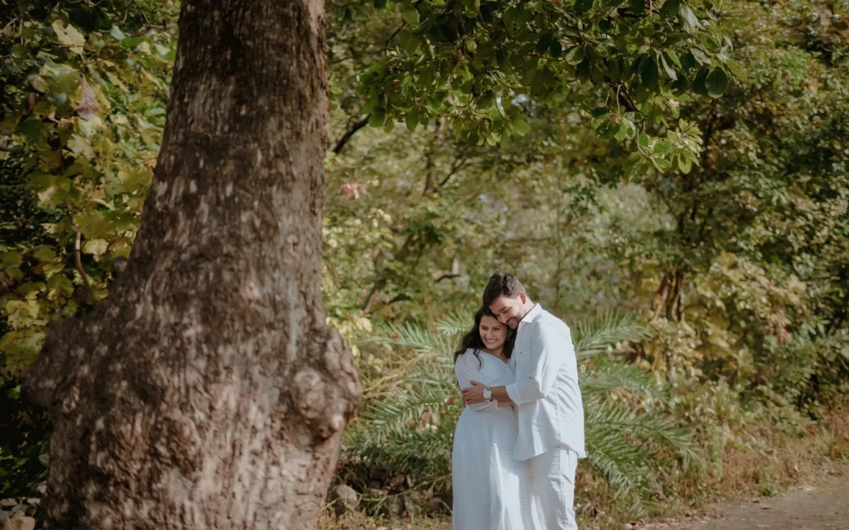 Couple in pale outfits embracing beside a large tree in a leafy outdoor setting