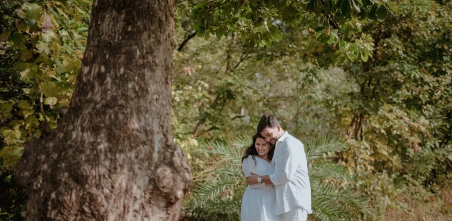 Couple in pale outfits embracing beside a large tree in a leafy outdoor setting