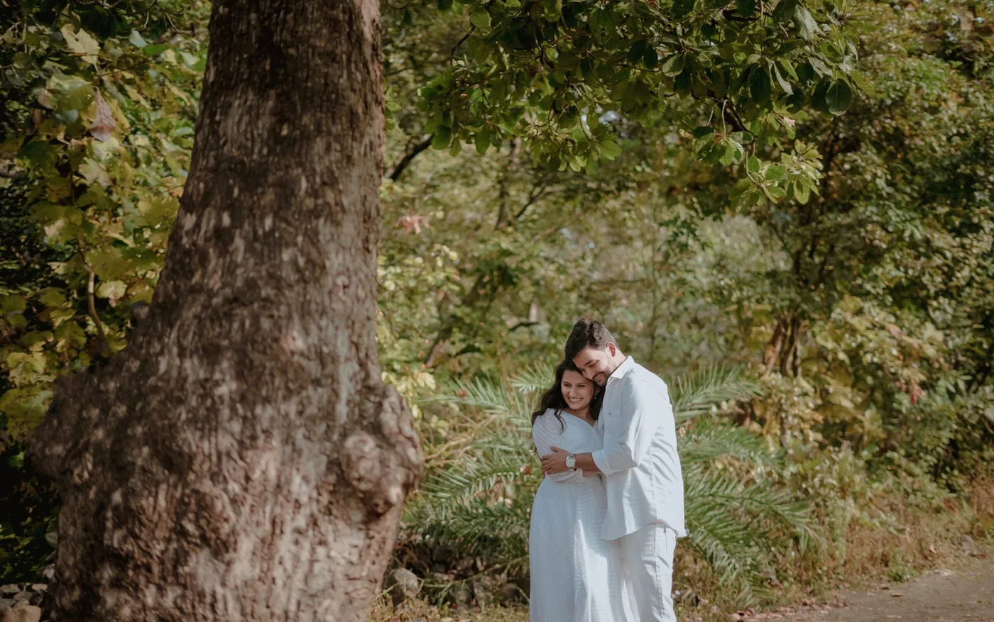 Couple in pale outfits embracing beside a large tree in a leafy outdoor setting