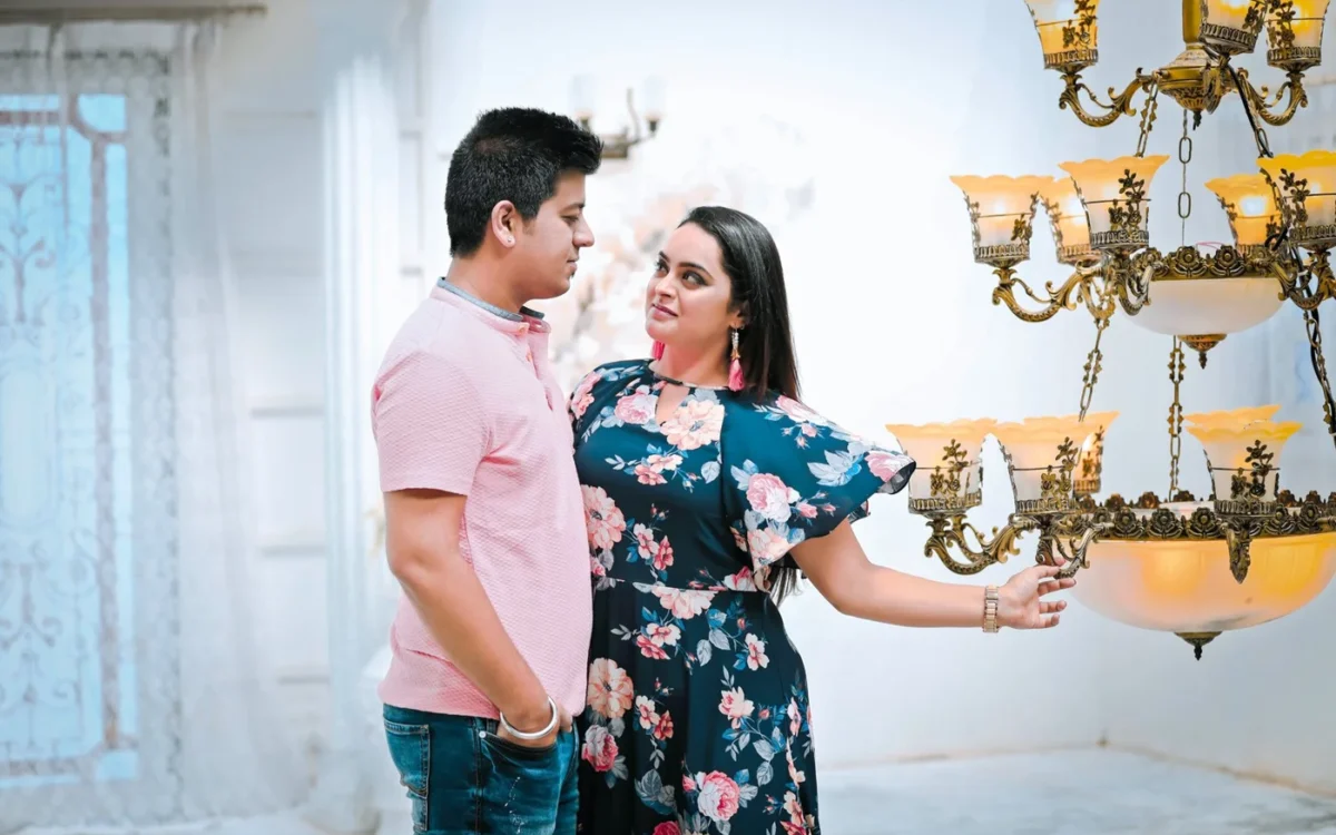 Man in a pink shirt and woman in a floral dress looking at each other beside a gold chandelier, a relaxed candid-style couple portrait in Indore