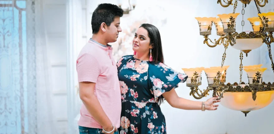 Man in a pink shirt and woman in a floral dress looking at each other beside a gold chandelier, a relaxed candid-style couple portrait in Indore