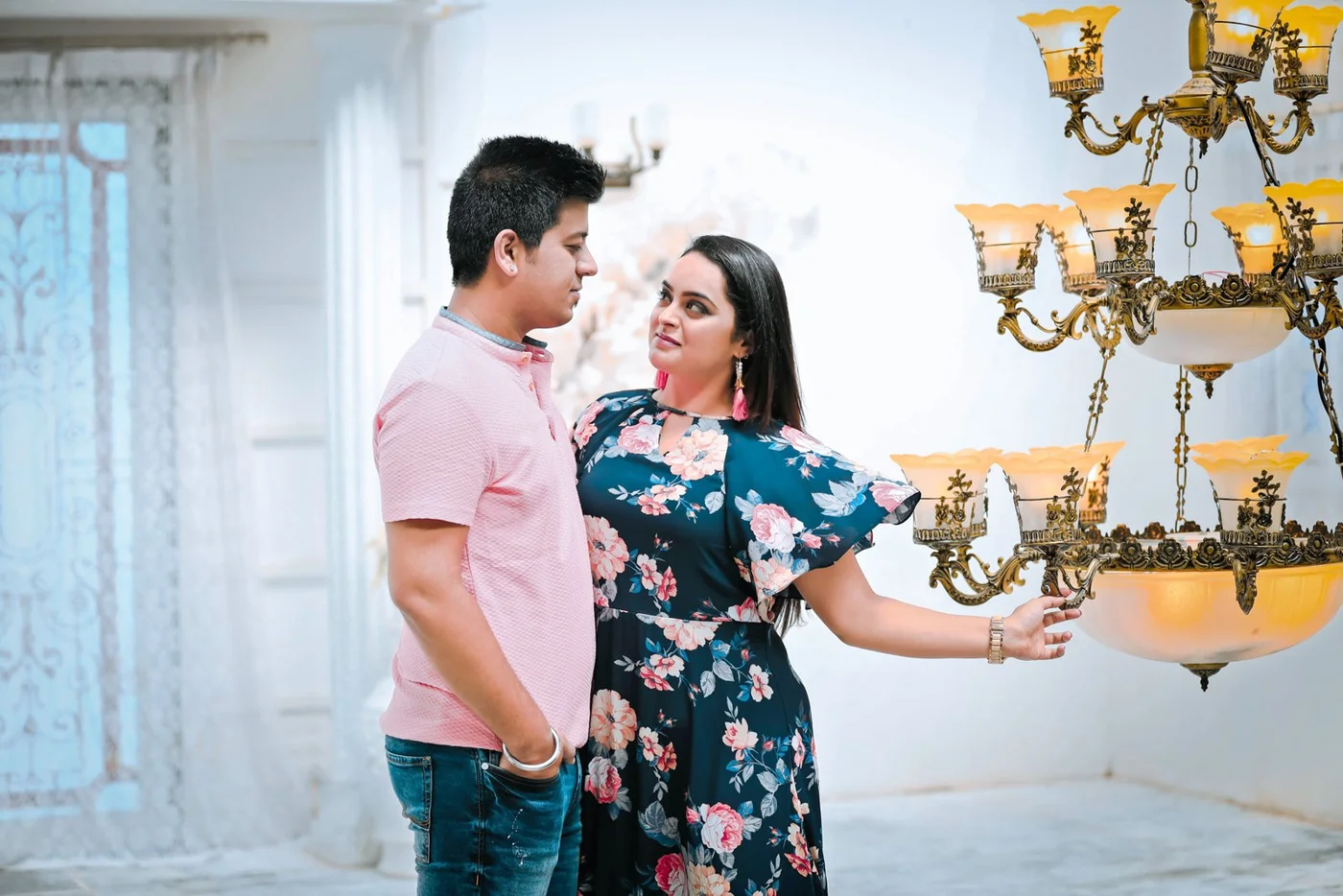 Man in a pink shirt and woman in a floral dress looking at each other beside a gold chandelier, a relaxed candid-style couple portrait in Indore