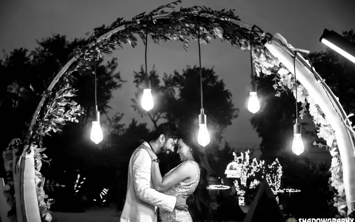Couple embracing under hanging bulbs and fairy lights in black and white, evening pre-wedding shoot