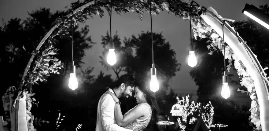 Couple embracing under hanging bulbs and fairy lights in black and white, evening pre-wedding shoot
