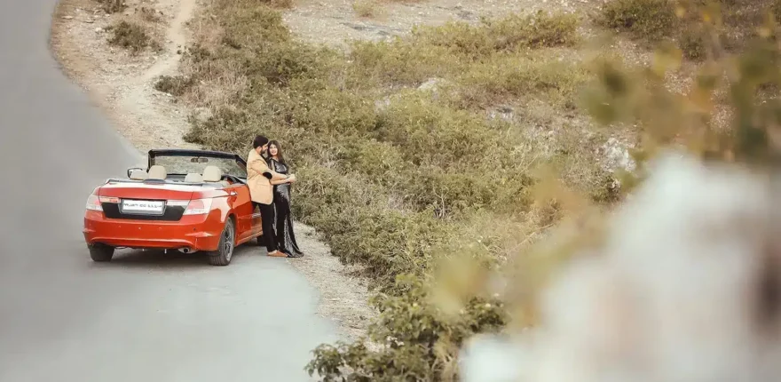 Couple standing beside a red car on a rocky hillside road, pre-wedding shoot