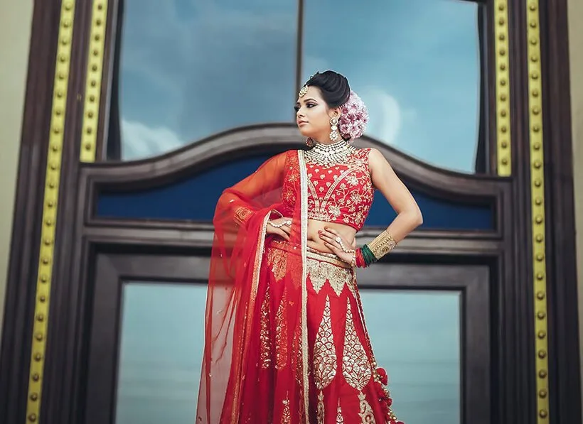 Bride in a red lehenga standing in front of a tall window, wedding portrait