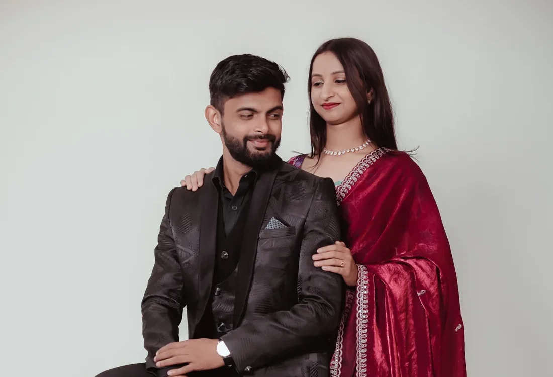 Groom in a black suit seated on a wooden stool with the bride in a red saree standing beside him in a studio portrait