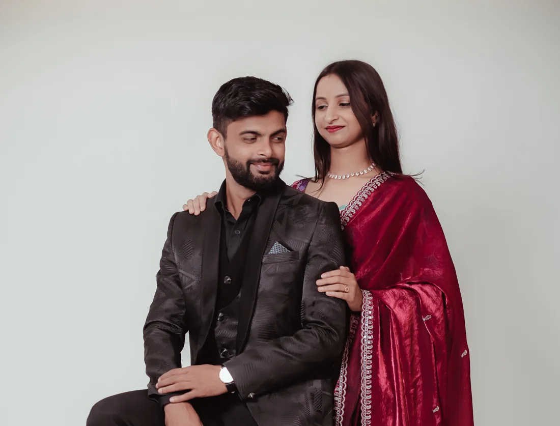 Groom in a black suit seated on a wooden stool with the bride in a red saree standing beside him in a studio portrait